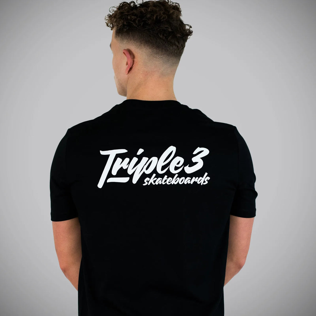 A man viewed from behind wearing a black t-shirt with a white 'Triple 3 skateboards' logo printed across the back, set against a plain gray studio background.