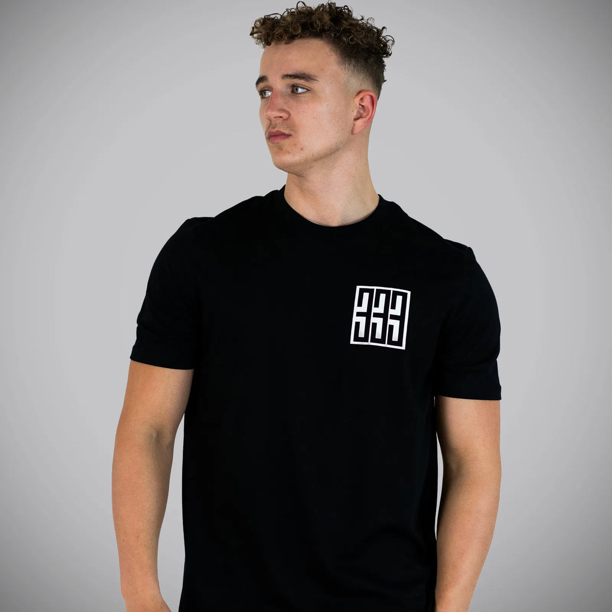 A man wearing a black short-sleeve t-shirt with a white geometric logo on the left chest, photographed against a neutral gray background.