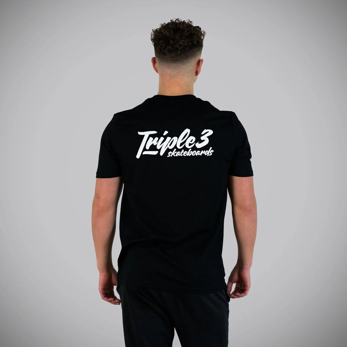 A man viewed from behind wearing a black t-shirt with a white 'Triple 3 skateboards' logo printed on the back, set against a plain gray studio background.