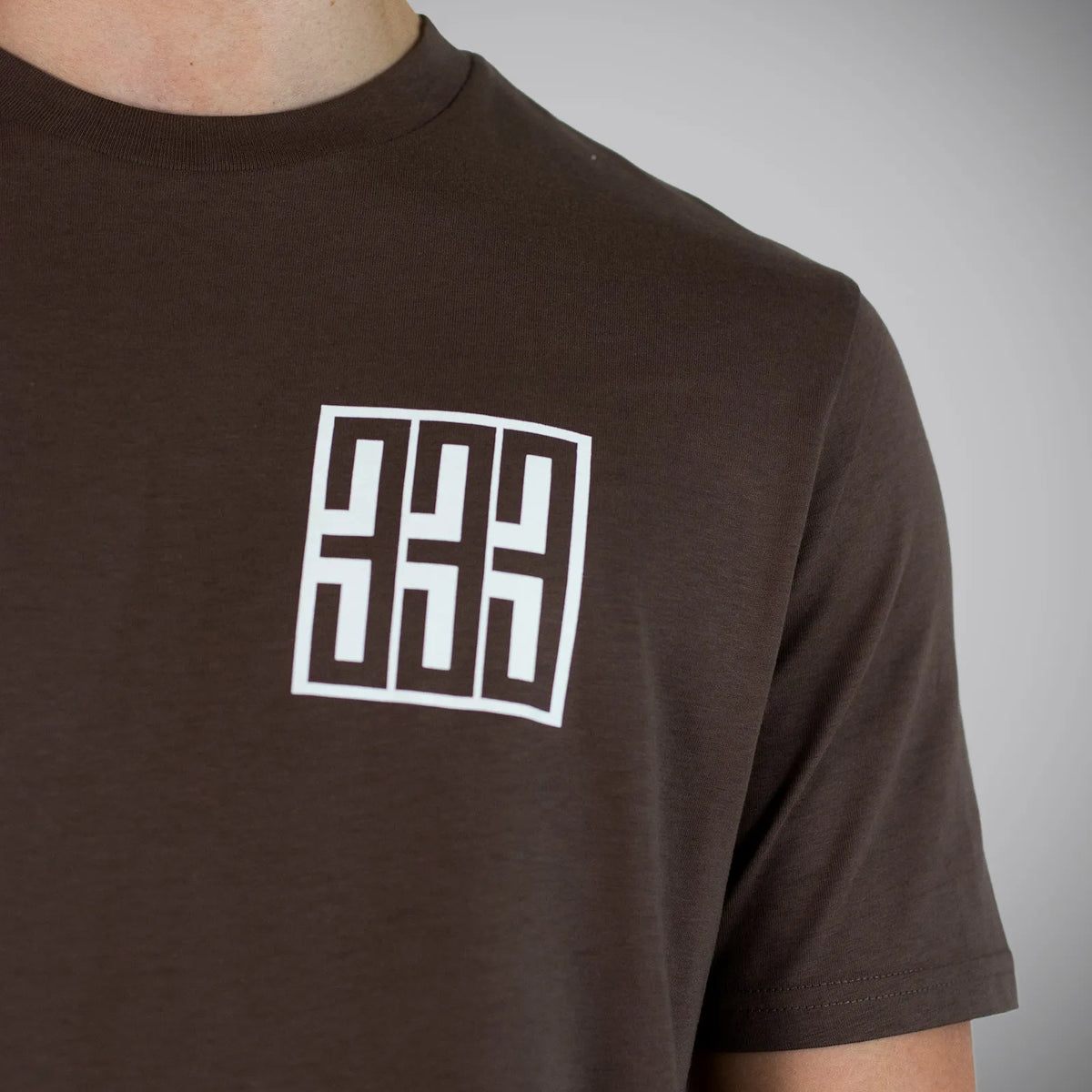 A close-up of a person wearing a dark brown crew-neck t-shirt with a white geometric logo printed on the chest, set against a neutral gray background.