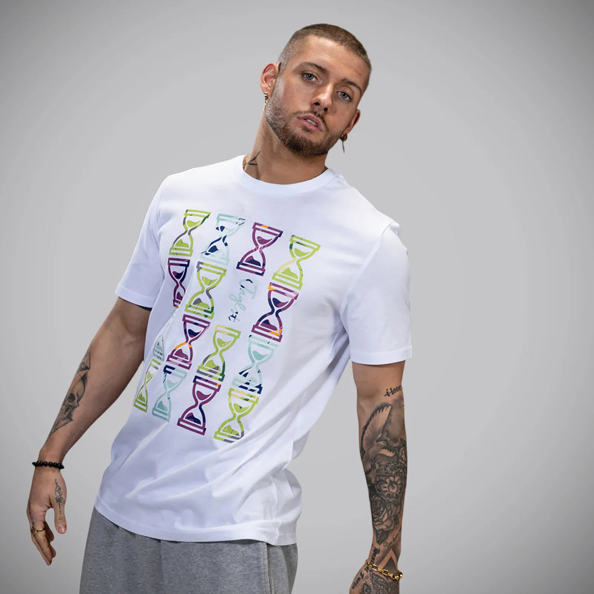 A man wearing a white short-sleeve t-shirt with a colorful graphic print of hourglasses, paired with gray sweatpants, against a solid gray studio background.