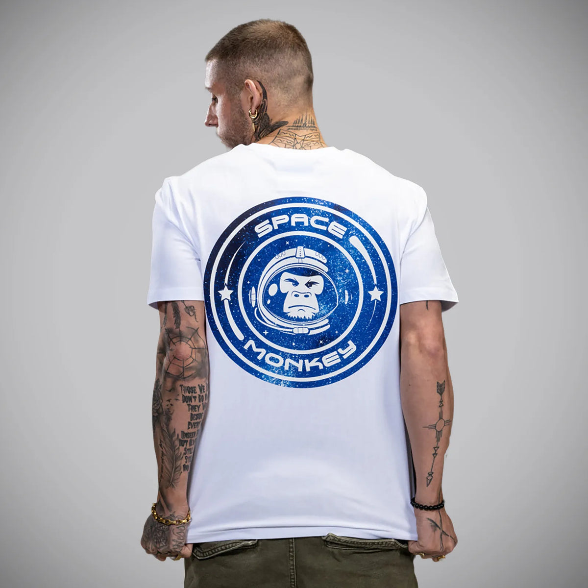A man viewed from behind wearing a white t-shirt with a large blue circular 'SPACE MONKEY' graphic on the back, paired with olive green pants, against a neutral gray studio background.