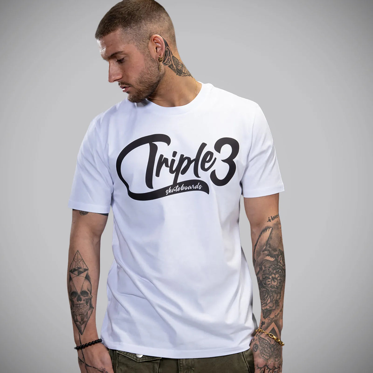 A man wearing a white crewneck t-shirt with a black 'Triple3 skateboards' graphic on the chest, photographed against a neutral gray background.