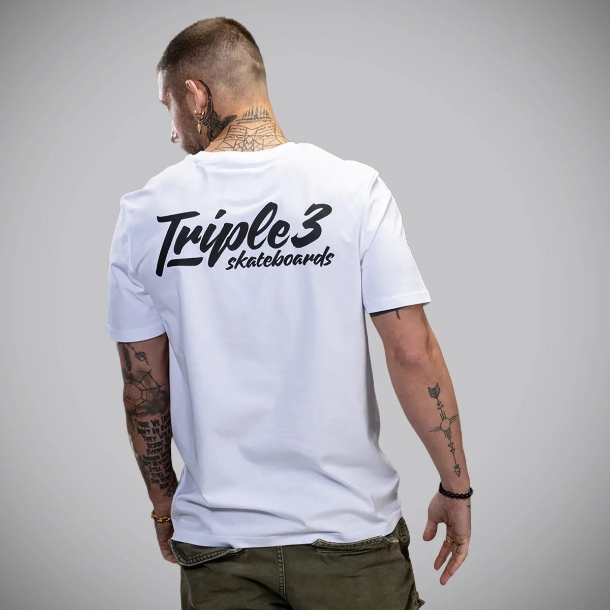 A man viewed from behind wearing a white short-sleeve t-shirt with 'Triple 3 skateboards' printed in black across the back, paired with olive green pants, against a solid gray studio background.